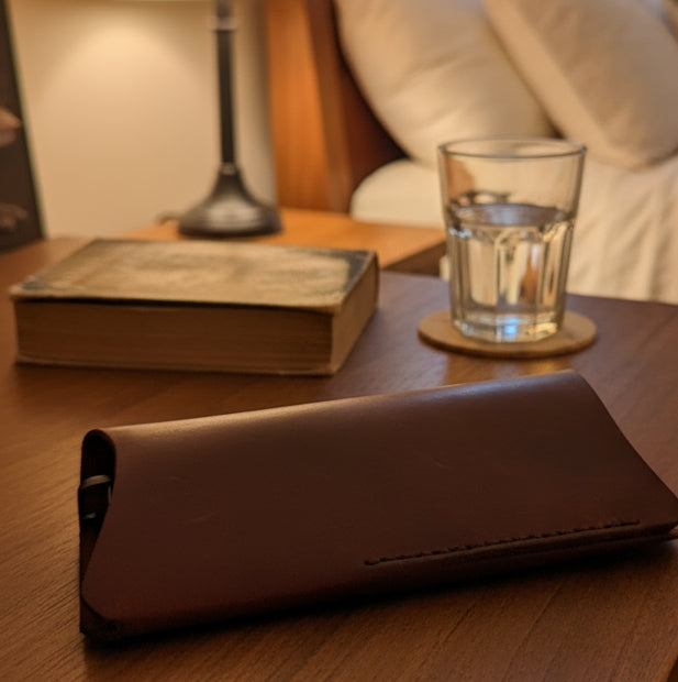 Hand sewn waxed leather glasses case on a wooden table next to a glass of water and a book.